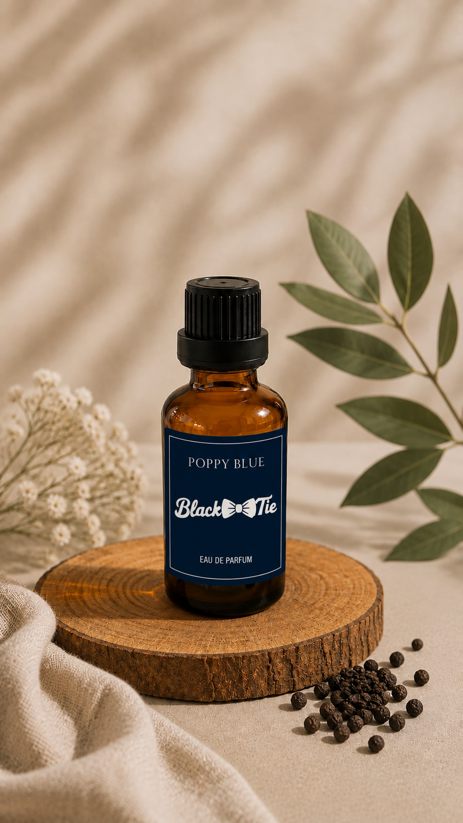 A small amber glass perfume bottle with a black cap and a dark blue label reading 'Poppy Blue' and 'Black Tie' stands on a round wooden slice. The scene is styled with beige fabric, scattered peppercorns, dried flowers, and green leaves against a softly lit neutral background.